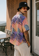 Man wearing a colorful shirt and black bucket hat standing on a porch.
