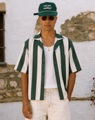 Person wearing a green and white striped shirt, white pants, and a green cap with text, standing against a textured wall.