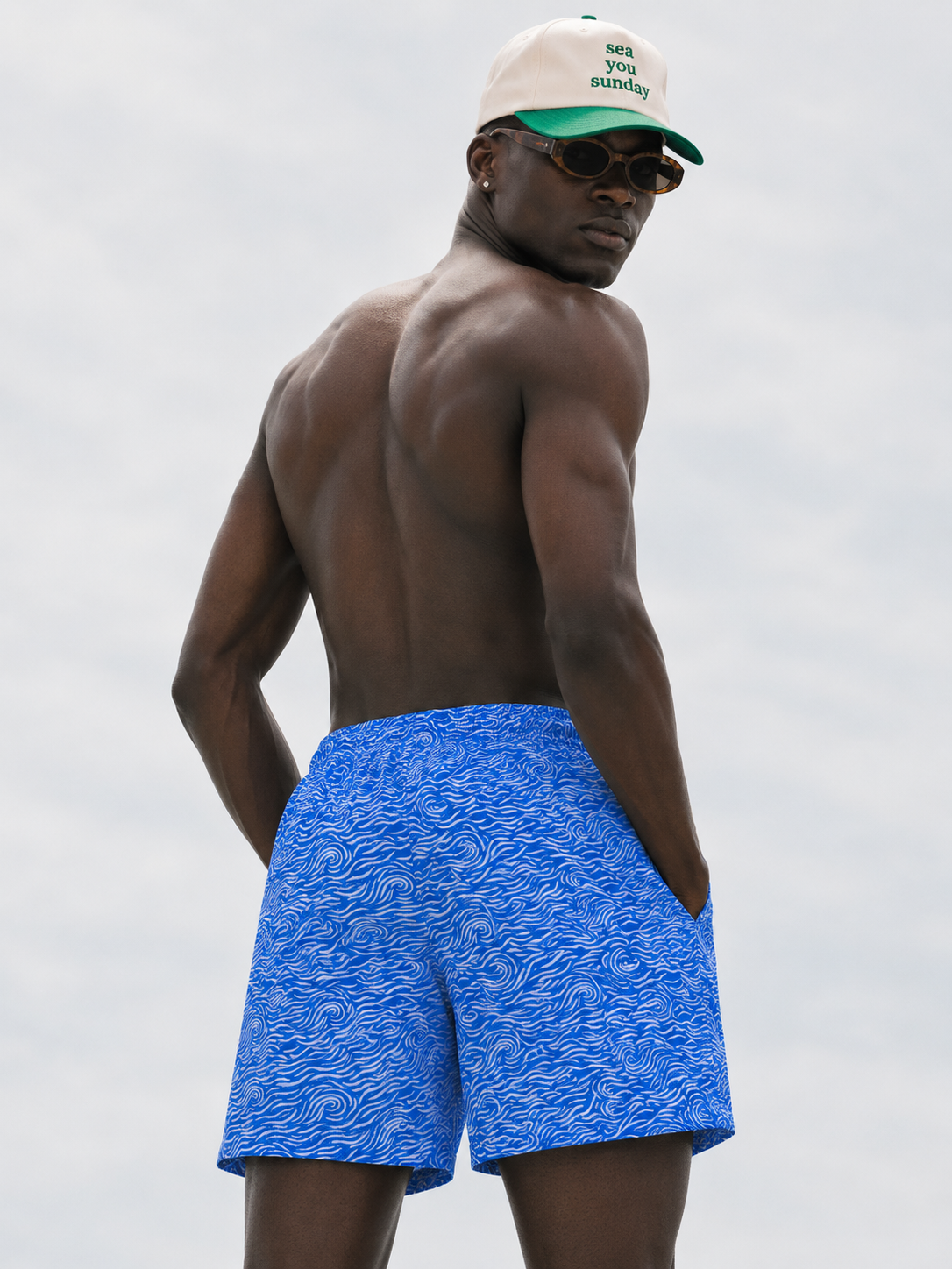 Man wearing blue swim shorts and a cap with sunglasses, standing against a plain background