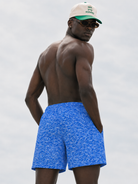 Man wearing blue swim shorts and a cap with sunglasses, standing against a plain background