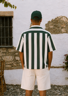 Man wearing a green and white striped shirt and cap standing in front of a white wall with floral decorations.