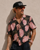 Man wearing a black cap with 'See You Sunday' text, sunglasses, and a pink floral shirt against a beige wall.