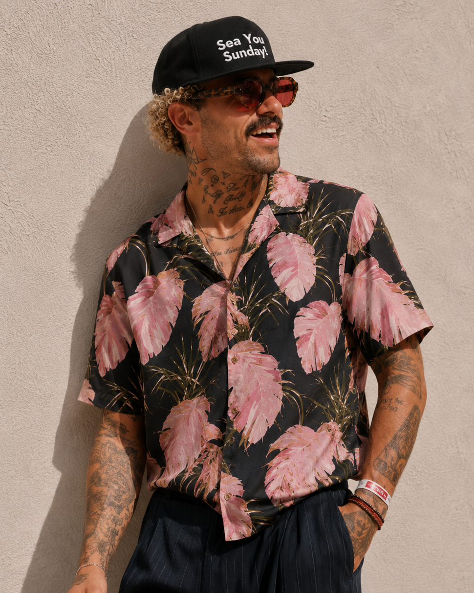 Man wearing a black cap with 'See You Sunday' text, sunglasses, and a pink floral shirt against a beige wall.