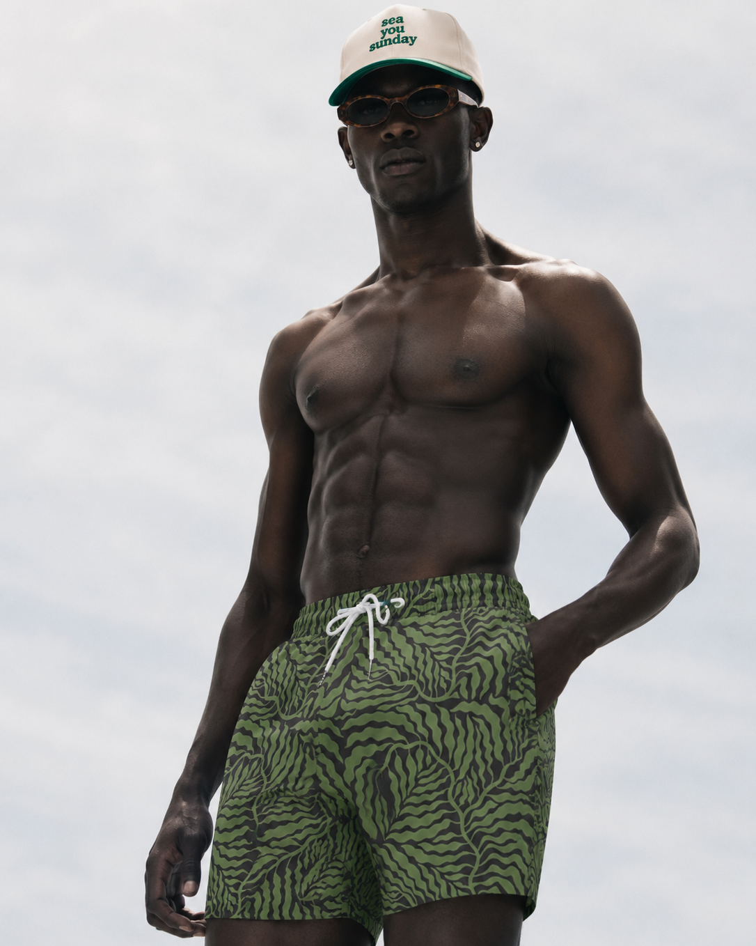 Man wearing green leaf-patterned shorts and a cap against a light gray background