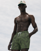 Man wearing green leaf-patterned shorts and a cap against a light gray background