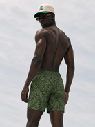 Man wearing green patterned swim shorts and a beige cap against a light gray background