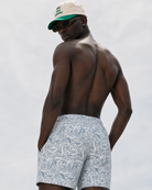 Man wearing patterned swim shorts and a cap on a plain background