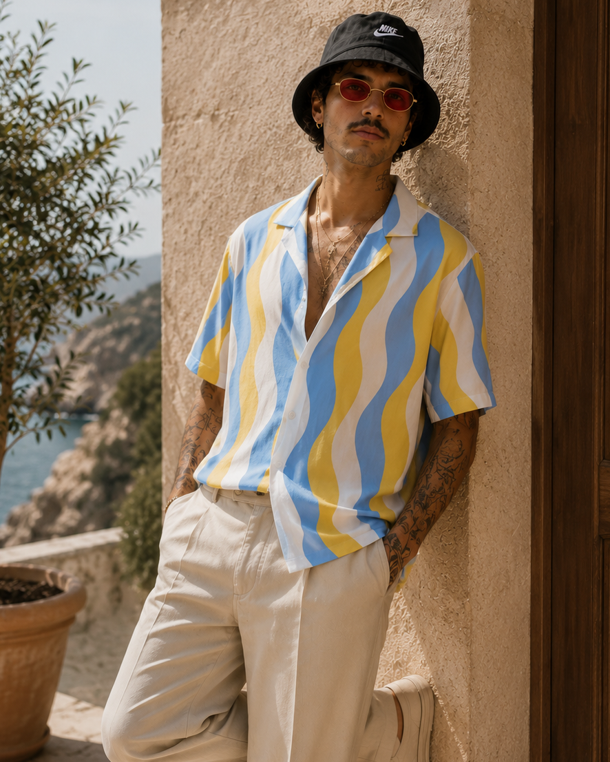Man wearing a colorful striped shirt and bucket hat leaning against a wall with a scenic background.